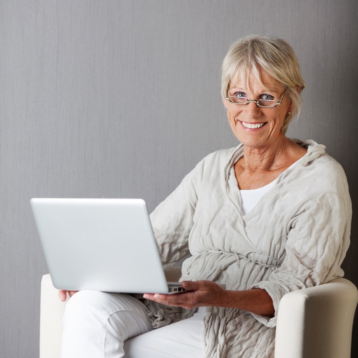 Portrait of happy senior woman with laptop sitting on couch against grey wall